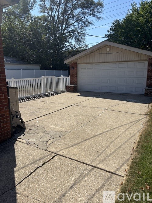 A driveway leads to a garage with a white fence and trees in the background.