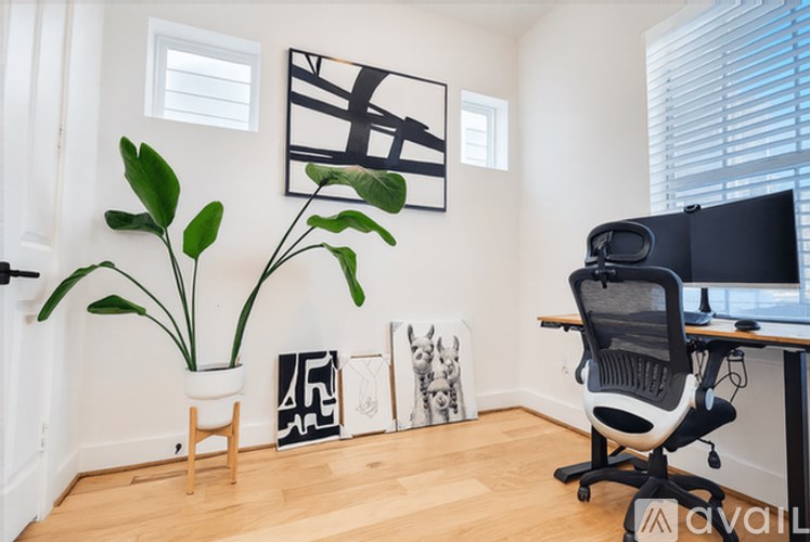 A room with a black and white picture on the wall, a potted plant, and a computer desk with a chair.