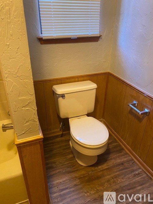 A white toilet in a small bathroom with wood paneling and a window.