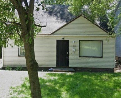 A small house with a black door and a window is surrounded by green grass and trees.