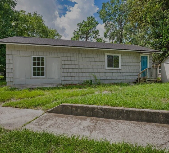 A small house with a blue door and a window is surrounded by grass and trees.