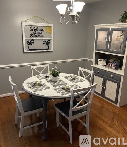 A dining room with a white table and chairs, a framed sign that says "Welcome to our Paradise", and a cabinet with a WI-FI box on top.