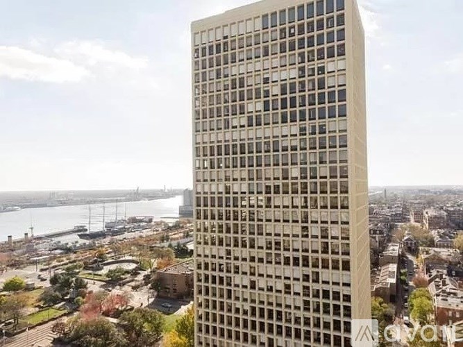 A tall building with a grid of windows stands in the foreground with a cityscape and a body of water in the background.