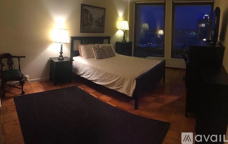 A bedroom with a bed, a chair, and a window overlooking a cityscape at night.