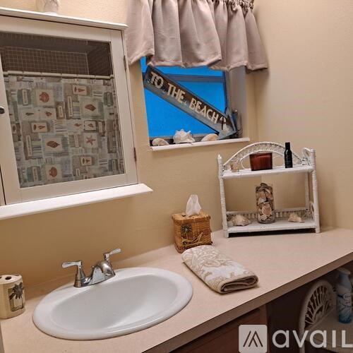 A bathroom with a white sink and a window with a curtain that says "To the Beach".