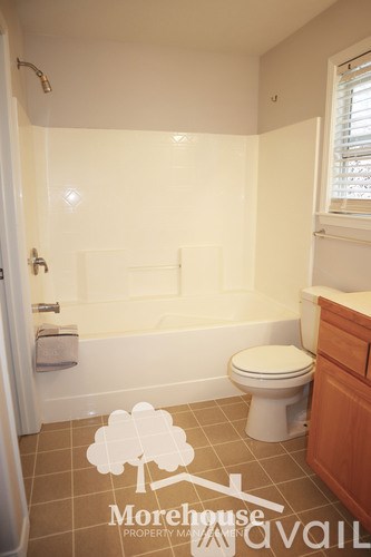 A white toilet sits in a bathroom with a white tub and a window.
