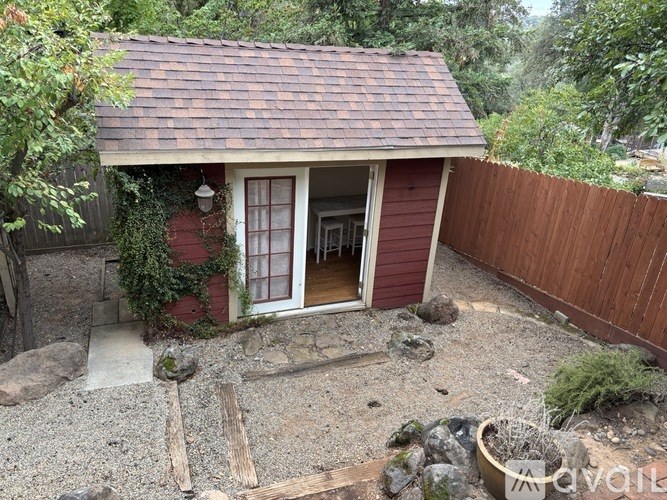 A small red house with a brown roof and a white door is surrounded by a gravel area and a wooden fence.