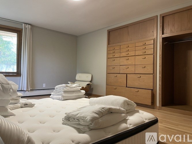 A white bed with pillows and a stack of towels on top of it in a room with wooden drawers and a window.