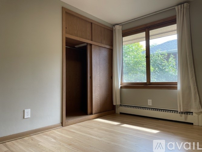 A room with a wooden floor, a window with white curtains, and a wall-mounted air conditioner unit.