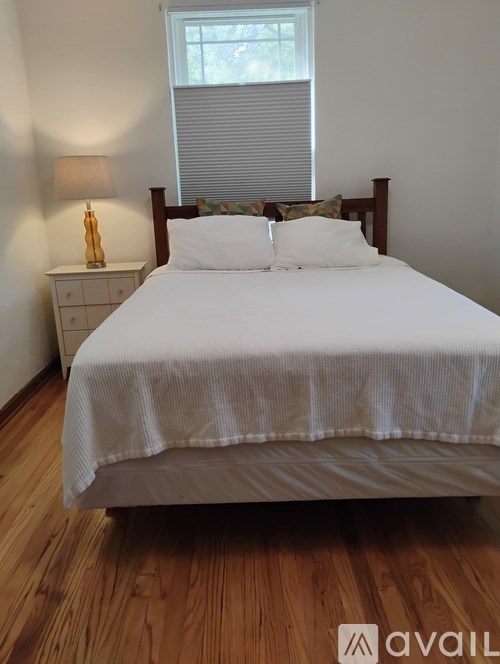 A bed with a white comforter and two pillows sits in a room with a wooden floor and a window with blinds.