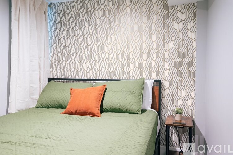 A bed with a green cover and two pillows, one orange, sits in a room with a patterned wallpaper.