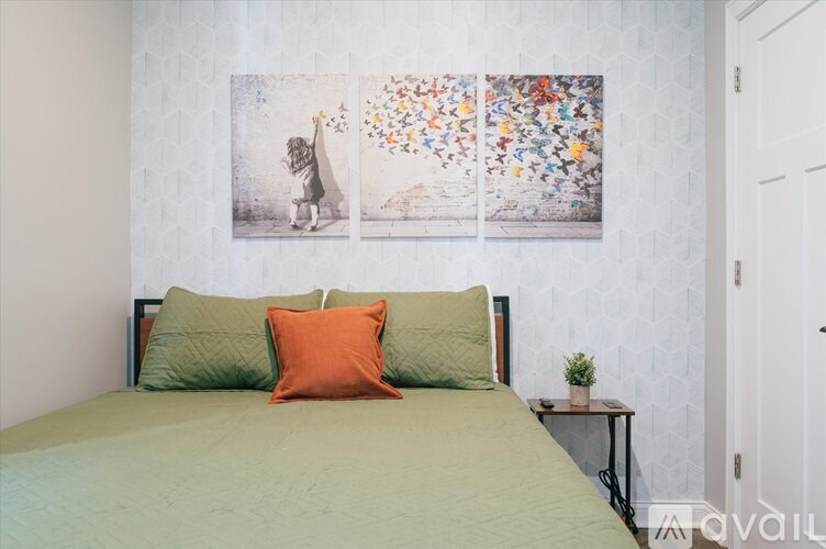 A bedroom with a bed, two pillows, a small table with a plant, and three pictures on the wall.