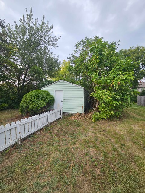 A green shed sits in a grassy yard next to a white picket fence.