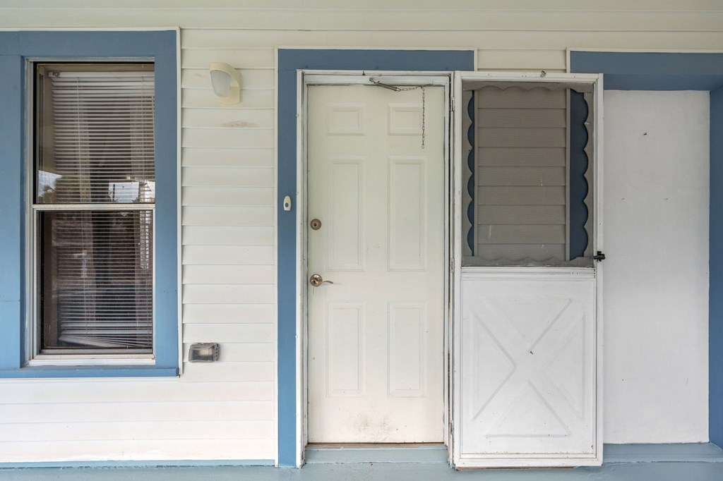 A white door is open to a room with a white wall and a window with blinds.
