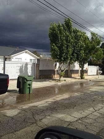 A car is parked on a street with a green trash can and a white house in the background.
