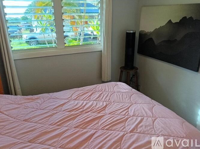 A bed with a pink comforter sits in a room with a window and a painting on the wall.