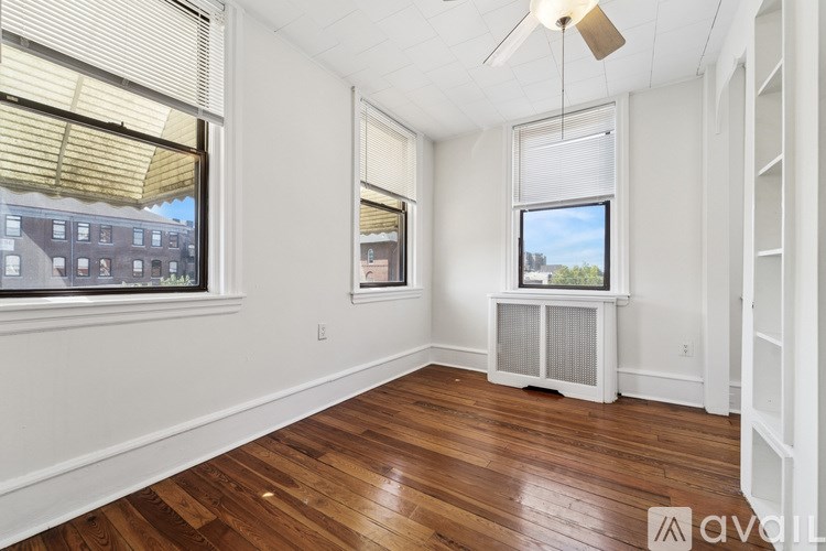 A room with wooden floors and white walls, featuring a window with blinds and a ceiling fan.