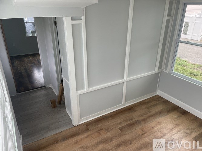 A room with white walls and wooden flooring, with a window showing greenery outside.