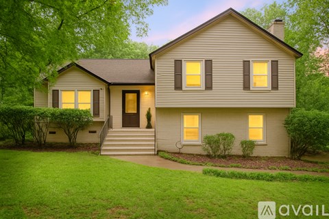 A house with a brown door and two windows is surrounded by greenery.