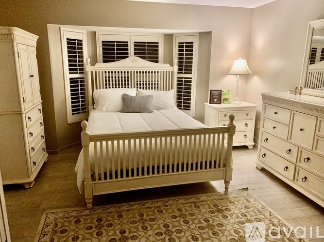 A white bed with a white headboard and pillow is in the center of a room with a white dresser and nightstand.