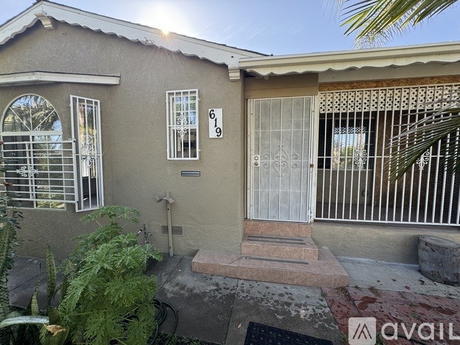 A house with a brown roof and a white door is surrounded by green plants.