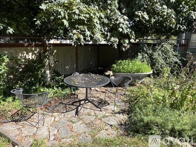 A patio table with chairs and a grill is surrounded by greenery.