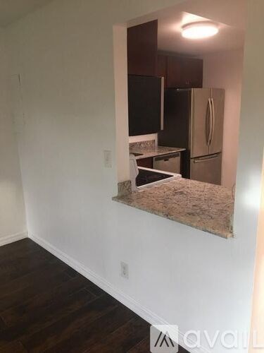 A kitchen with a refrigerator and cabinets is visible through a window in a room with white walls and wood flooring.