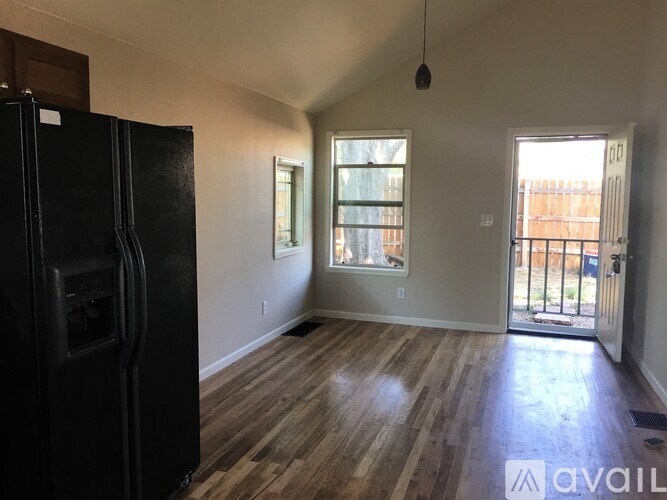 A black fridge stands in a room with wooden floors and two windows.
