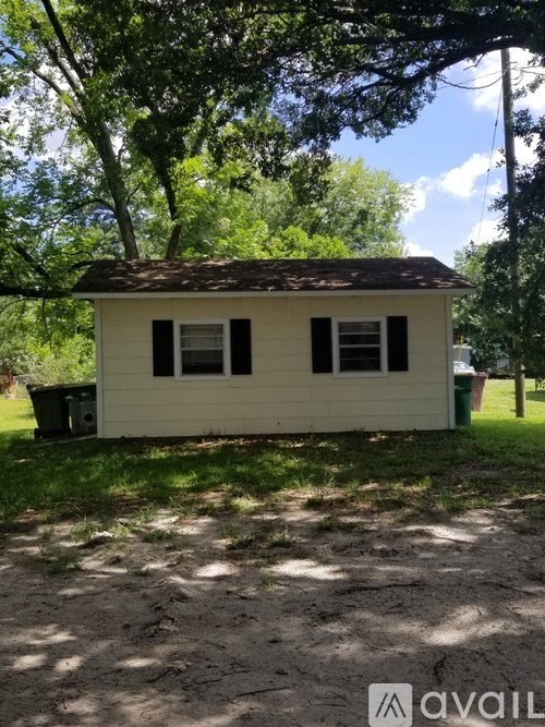 A small white house with a brown roof is situated in a grassy area with trees in the background.