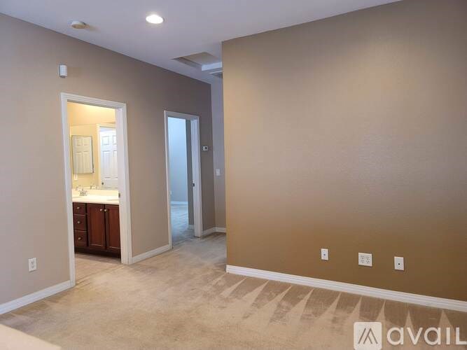 A room with beige walls and carpeting, a door, and a small kitchen area with a sink and cabinets.