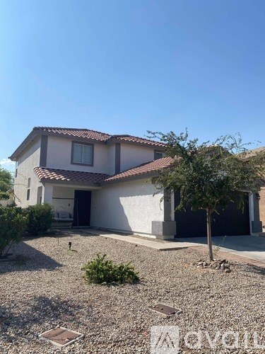 A house with a white exterior and a brown roof is surrounded by a gravel driveway and a small tree.