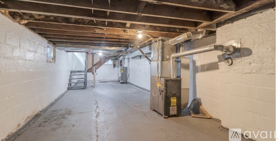 A room with a concrete floor and walls, featuring a ladder, a metal box, and a pipe.