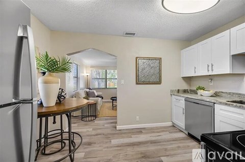 A kitchen with a refrigerator, a table with a plant on it, and a view of a living room through a window.
