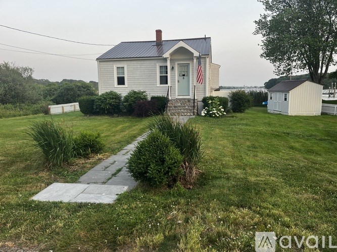 A house with a flag on the front porch is surrounded by a grassy lawn and a small garden.