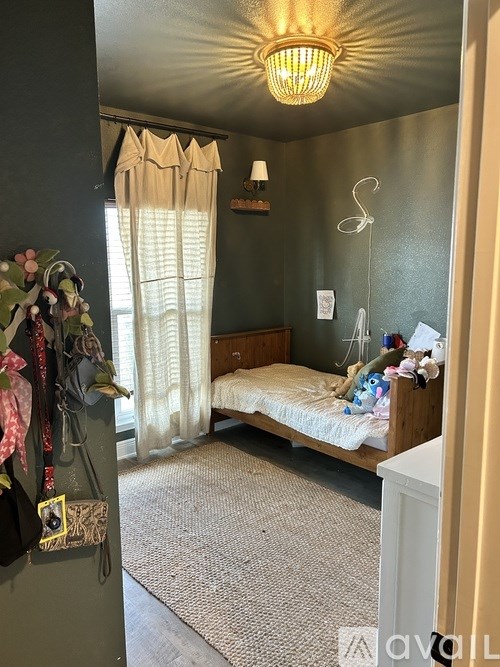 A small bedroom with a bed, a window with curtains, a hanging chair, and a wall-mounted shelf with various items.