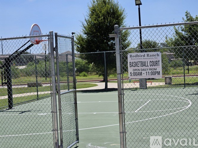 A basketball court is surrounded by a fence and has a sign that reads "Redbird Ranch BASKETBALL COURT OPEN DAILY FROM 5:00AM - 11:00PM".
