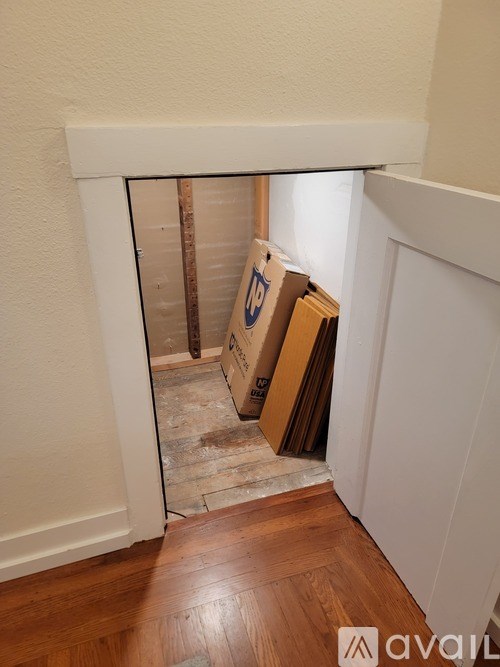 A room with a white wall and a wooden floor with a cardboard box and a wooden plank.