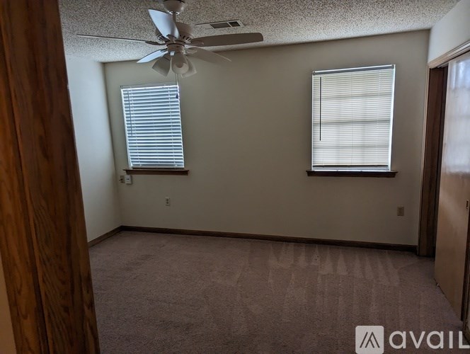 A room with a carpeted floor, a ceiling fan, and two windows with blinds.