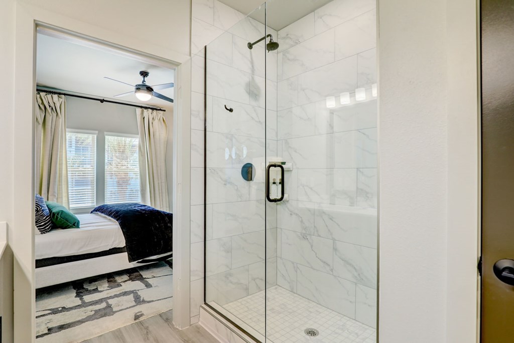 A bathroom with a walk-in shower and a bedroom with a bed and a ceiling fan.
