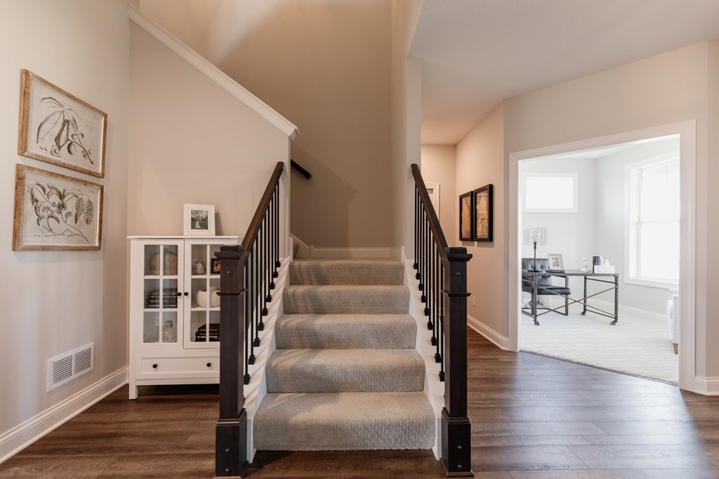 A staircase with a black railing and a grey carpeted runner leads to a bright room with a table and chairs.