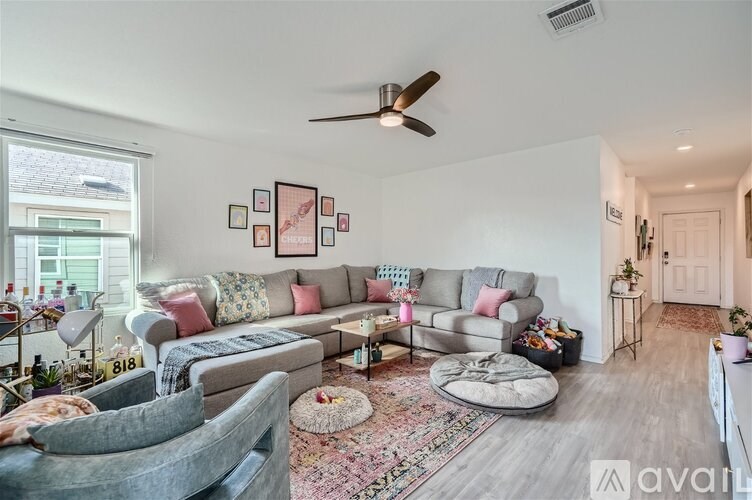 A living room with a grey couch, a grey armchair, a grey ottoman, a rug, a coffee table, a ceiling fan, a wall with pictures, a door, a plant, and a window.