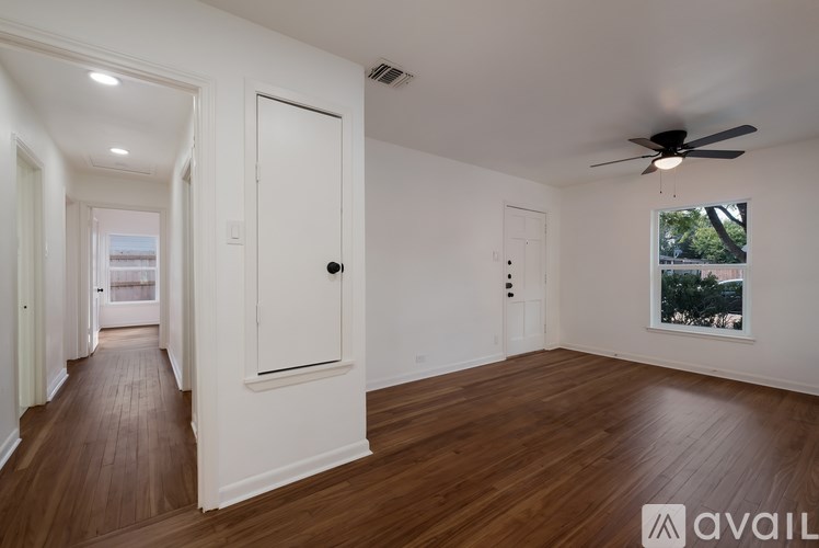 A spacious room with wooden floors and white walls, featuring a ceiling fan and a window with a view of trees outside.