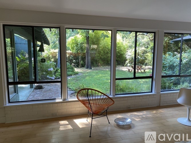 A room with a chair and a table in front of a window with a view of a garden.