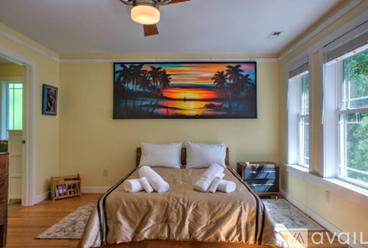 A bedroom with a bed, a ceiling fan, and a painting of a sunset on the wall.
