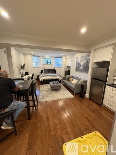 A man is sitting at a desk in a room with wooden floors and a bed in the background.