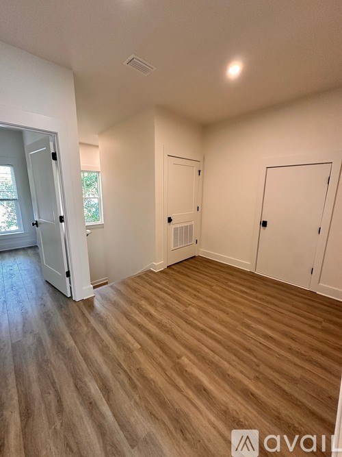 A room with wooden flooring and white walls, with a window on the left and a door on the right.
