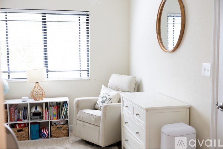 A white chair with a pillow is in the corner of a room with a white wall and a round mirror.