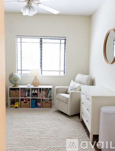 A room with a white chair, a bookshelf, a window with blinds, a ceiling fan, and a round mirror on the wall.