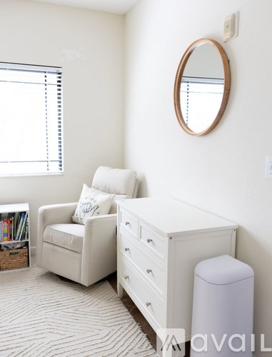 A white chair with a pillow is in front of a white wall with a round mirror above it.