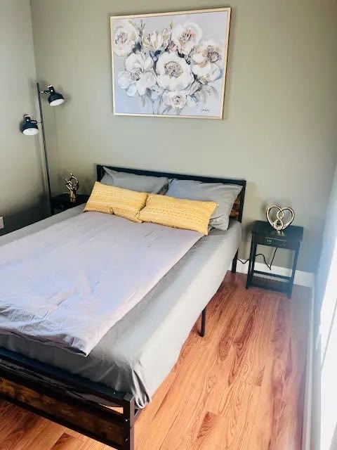 A bed with a white sheet and two yellow pillows sits in a room with a floral picture on the wall.